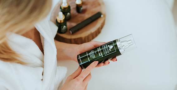 Person i vit morgonrock h&aring;ller en gr&ouml;n flaska med hudv&aring;rdsprodukt, med fler flaskor i bakgrunden p&aring; en tr&auml;bricka.