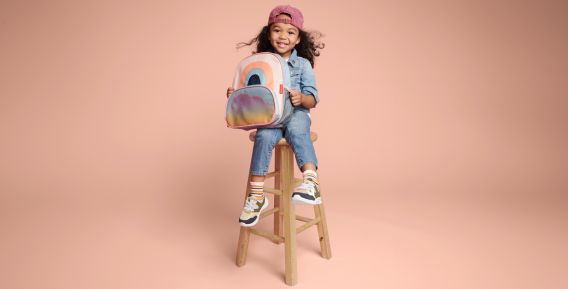 Ett leende barn sitter p&aring; en h&ouml;g tr&auml;pall mot en beige bakgrund. Barnet b&auml;r jeanskl&auml;der, en rosa keps och h&aring;ller i en f&auml;rgglad ryggs&auml;ck med regnb&aring;gsmotiv.