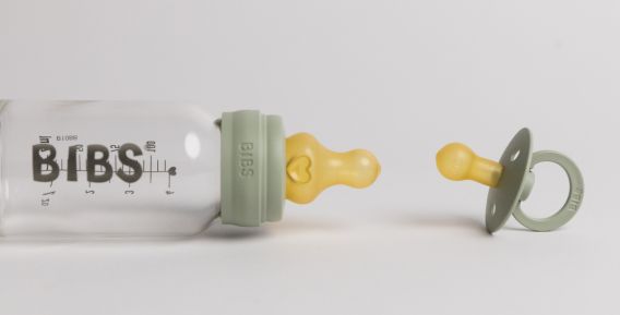 En BIBS-flaska med gul napp och en BIBS-napp med gr&aring; sk&ouml;ld ligger bredvid varandra p&aring; en vit bakgrund.
