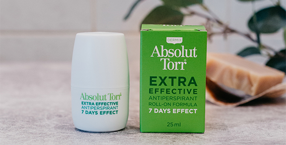 B&aring;de innerf&ouml;rpackning och ytterf&ouml;rpackning fr&aring;n Absolut Torr roll -on placerade bredvid varandra mot en gr&aring; b&auml;nkskiva med struktur. I bakgrunden syns en gr&ouml;n kvist och en fast tv&aring;l.