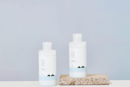 Tv&aring; RoundLab hudv&aring;rdsflaskor st&aring;r p&aring; ett korkblock framf&ouml;r en mjuk gradientbakgrund i bl&aring;tt och ljusrosa. Flaskorna har minimalistiska etiketter med bergsikoner och en ljusbl&aring; f&auml;rgdetalj.